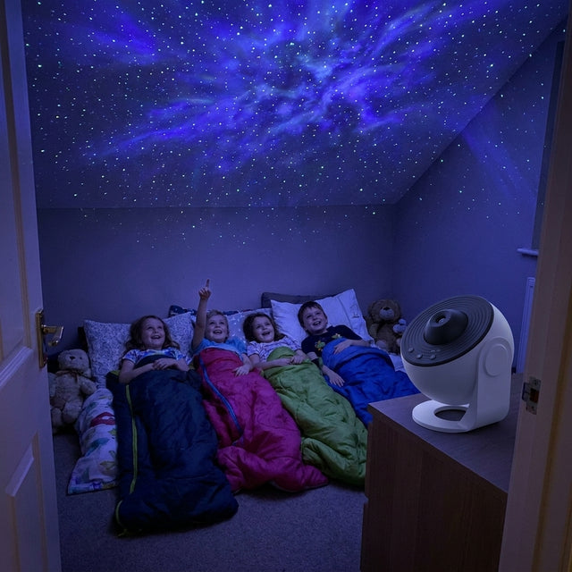 Four children in sleeping bags at a sleepover, looking up at a starfield and nebula projected across the whole bedroom ceiling