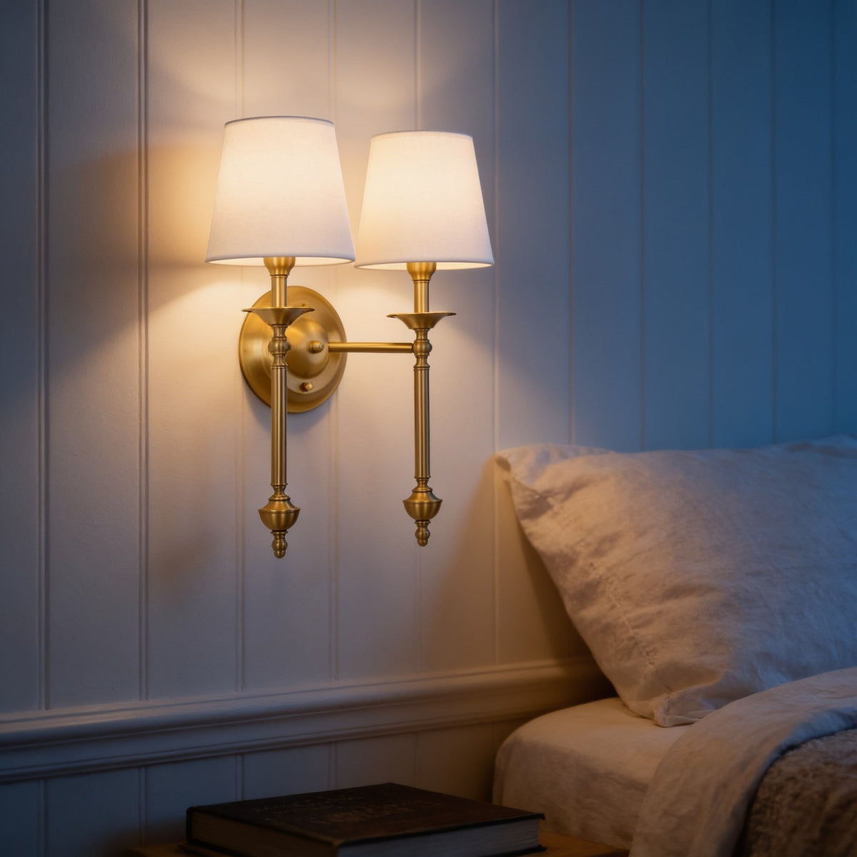Solid brass two-arm wall sconce with white fabric shades beside a bed