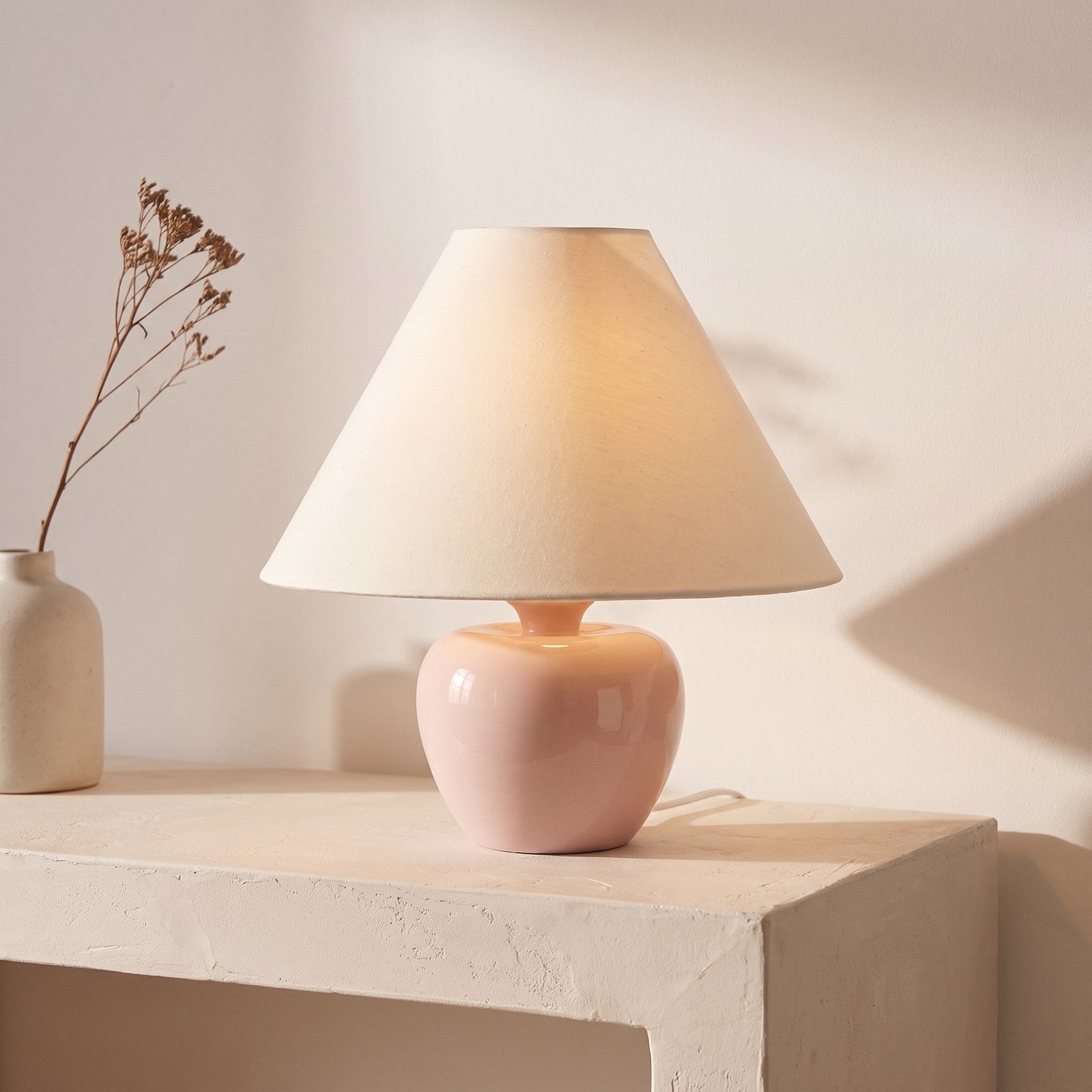 Blush pink apple-shaped ceramic table lamp with cream cone shade on a plaster console table