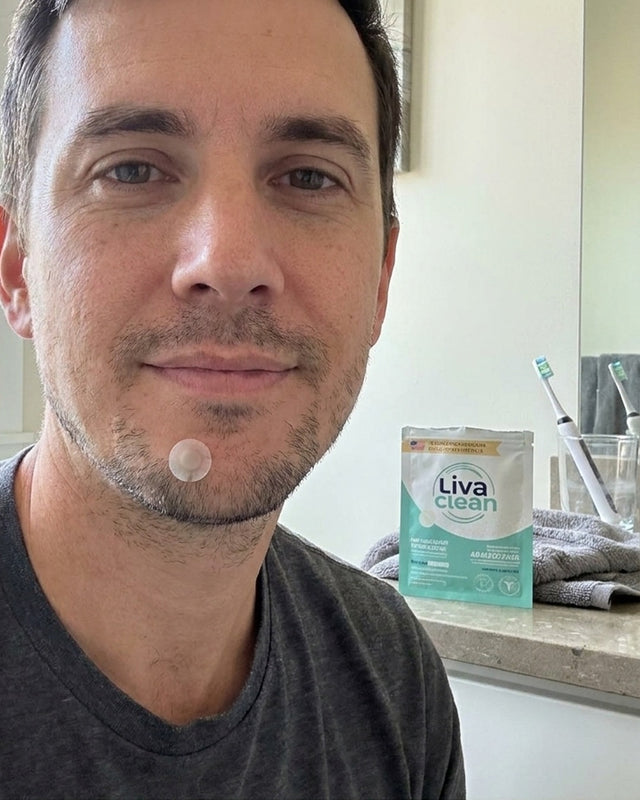 A man wearing a single hydrocolloid spot patch on his chin in a bright bathroom, LivaClean pouch on the counter