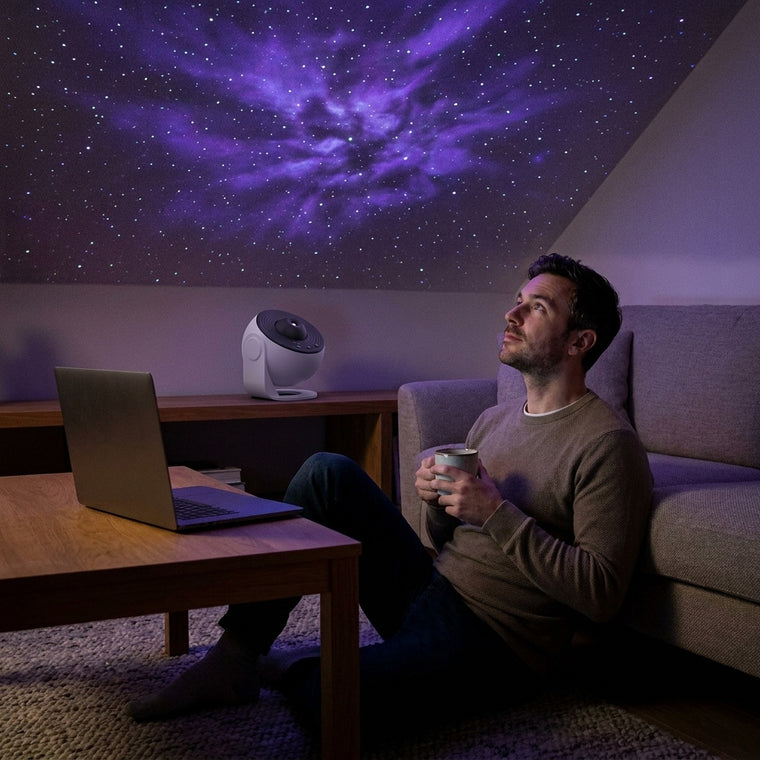Man sitting on his living room floor with a mug, laptop closed, looking up at a projected nebula